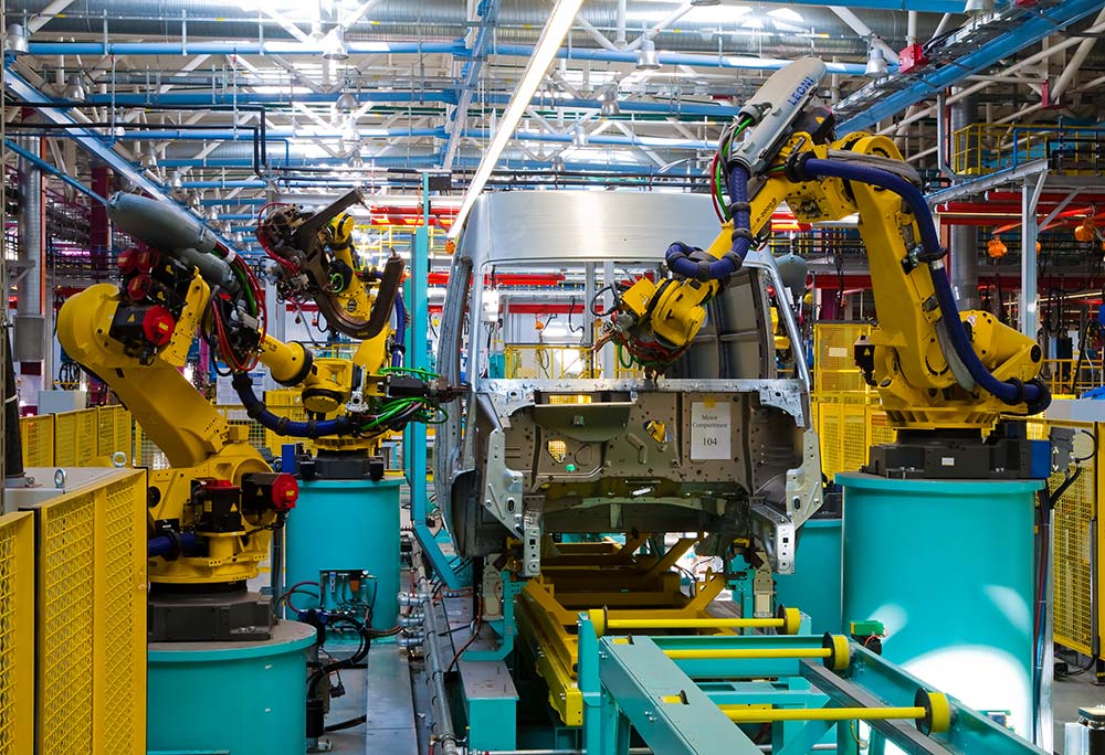 Two yellow robotic arms assemble a van as it moves down an automotive assembly line in a factory. 