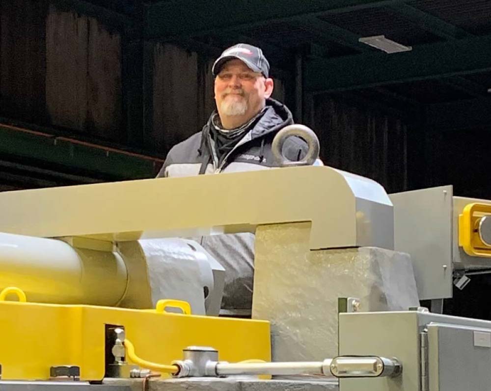 Male employee smiles while standing behind a piece of large equipment.