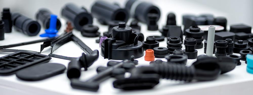 A wide range of mainly black rubber and plastics products for the automotive industry are arranged on a white tabletop.